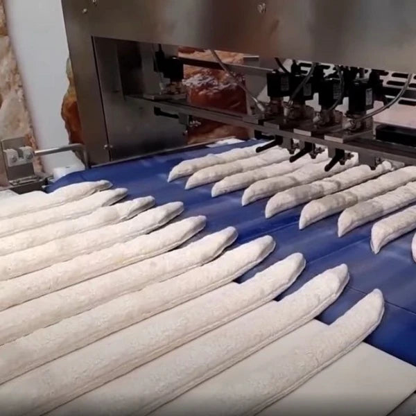 Long white bread loaves moving on a blue conveyor belt under a stainless steel industrial bakery machine.