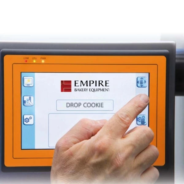 A close-up of a cookie depositor touchscreen control panel with an orange bezel and a hand touching the screen. The screen displays the Empire Bakery Equipment logo and interface icons.