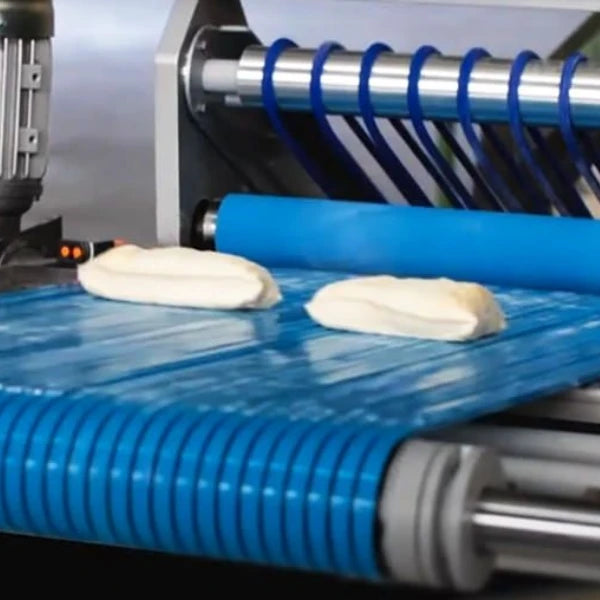 A close-up of a blue conveyor belt with two pieces of dough moving along the belt, with metal rollers and blue loops visible in the background.