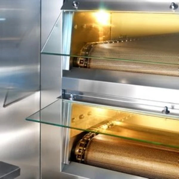 Close-up of stainless steel and glass oven doors on the tunnel oven with a warm glowing interior containing mesh rollers.