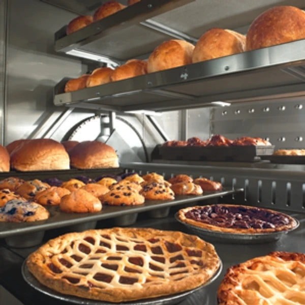 An inside look at revolving oven trays filled with freshly baked goods, including several large apple pies, assorted muffins, and rolls on metal shelves.